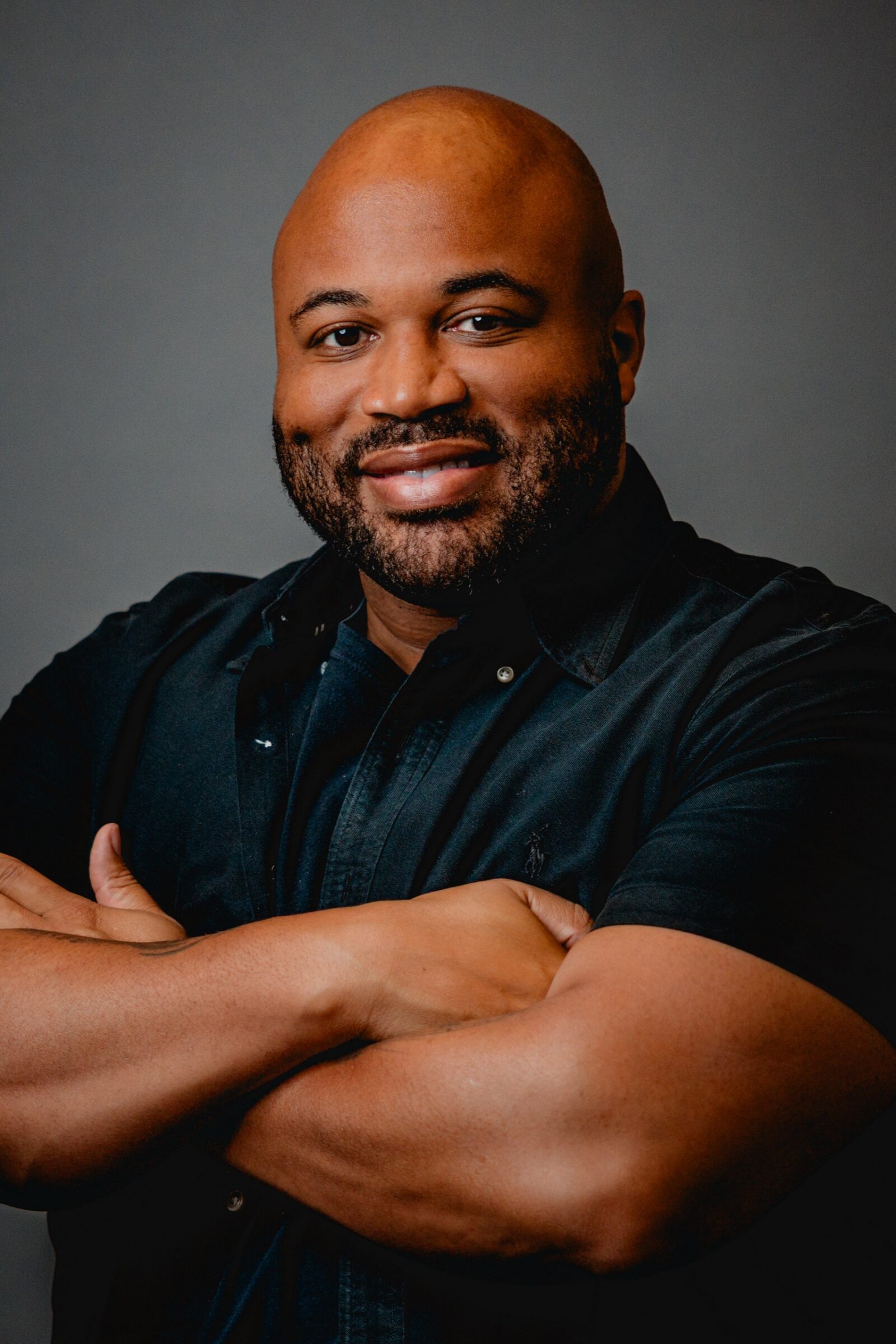 A smiling man in a dark shirt with his arms crossed, on a gray backdrop